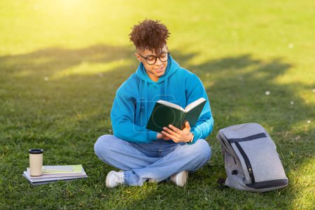 A focused young brazilian guy in blue hoodie sits cross-legged on the grass reading a book, with a backpack nearby
