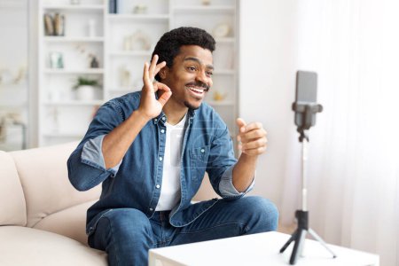 Téléchargez les photos : Un homme noir joyeux dans une chemise en denim gestuelle OK lors d'un appel vidéo via son smartphone sur un trépied, un homme afro-américain positif capturant du contenu à la maison - en image libre de droit