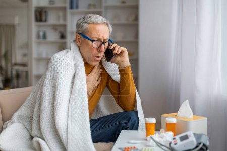 Photo for A senior man sits on a couch with a blanket over him, surrounded by medications and tissues, talking on the phone, depicting sickness - Royalty Free Image