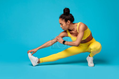 Concentrated African American sportswoman in vibrant yellow athleisure wear stretches during her workout on a blue backdrop