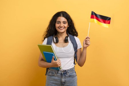 Foto de Una estudiante feliz con auriculares sostiene una bandera alemana y cuadernos coloridos, lo que sugiere el aprendizaje de idiomas extranjeros - Imagen libre de derechos