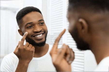 A joyful African american man applies face cream while smiling, reflecting a positive start to his morning routine