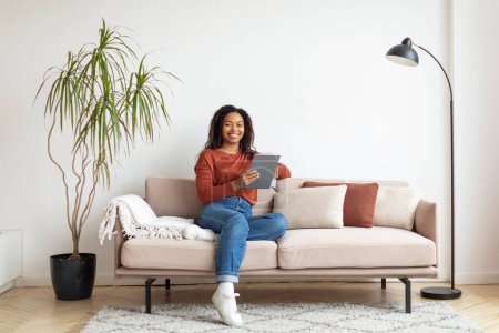 Foto de Mujer joven negro está sentado en un sofá, concentrándose en el uso de un dispositivo de tableta, mujer feliz relajarse con gadget moderno en casa - Imagen libre de derechos