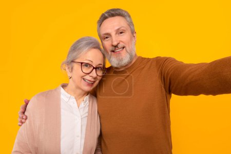 Téléchargez les photos : Portrait de vieil homme européen heureux embrassant sa femme dans des lunettes, posant pour selfie ensemble souriant à la caméra, debout sur fond de studio jaune. Médias sociaux et blogging, communication - en image libre de droit