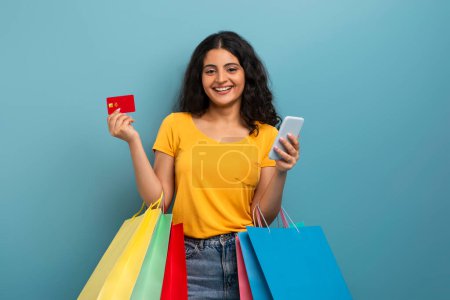 Eine fröhliche junge Frau lächelt, während sie eine Kreditkarte und ein Telefon vorzeigt, die die Leichtigkeit digitaler Transaktionen repräsentieren