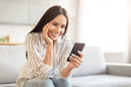 Téléchargez les photos : Femme au sourire contenté regardant son smartphone, assise sur un canapé lumineux et aéré du salon, espace de copie - en image libre de droit
