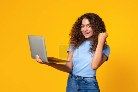 Foto de Imagen vibrante de una mujer sosteniendo un portátil y animando con una bomba de puño feliz en el fondo amarillo del estudio - Imagen libre de derechos
