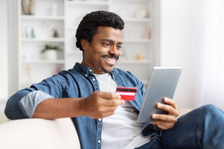 Ein fröhlicher schwarzer Mann, der eine Kreditkarte in der Hand hält, während er ein digitales Tablet benutzt, wahrscheinlich einen Online-Kauf von zu Hause aus tätigt und online einkauft