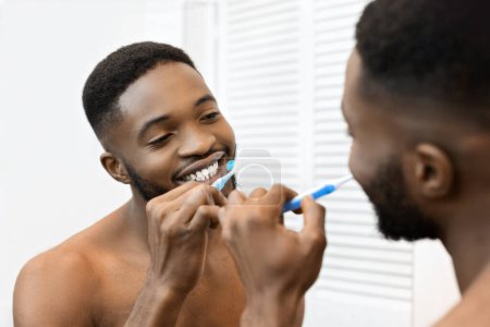 A shirtless African american man brushes his teeth in the mirror, representing good hygiene and personal care habits