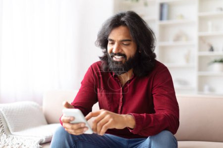 Lächelnder indischer Mann, der sich mit seinem Smartphone beschäftigt, bequem auf einem Sofa in einem angenehm beleuchteten Raum sitzend