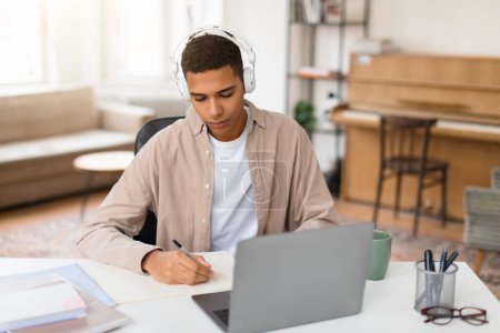 Téléchargez les photos : Focalisé jeune homme portant des écouteurs blancs écrit dans un carnet avec son ordinateur portable ouvert, engagé dans l'apprentissage - en image libre de droit