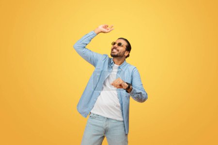 Photo for A carefree young indian man with a beard and sunglasses dances joyfully, wearing a light-blue shirt and jeans against a vibrant yellow background - Royalty Free Image