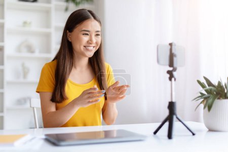 Eine fröhliche junge Asiatin im gelben Hemd nimmt mit einem Smartphone auf einem Stativ Inhalte für ihren Vlog auf