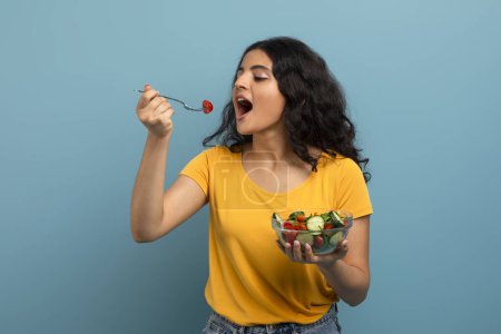 Junge Frau genießt eine Schüssel mit frischem Salat mit fröhlichem Gesichtsausdruck vor blauem Hintergrund