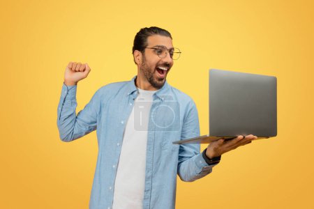 Elated indian man celebrating success or victory with an open laptop against a yellow backdrop