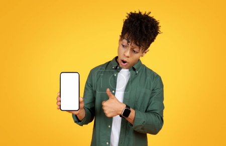 Téléchargez les photos : Jeune homme noir étonné montrant un écran de smartphone vide, exprimant son incrédulité, sur un fond jaune vibrant - en image libre de droit