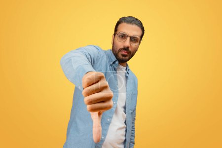 A bearded indian man in casual wear gives a thumbs down sign, showing disapproval or dissatisfaction, with a focused look, against a yellow background