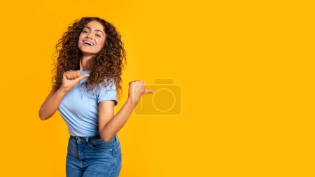 Photo for Young cheerful woman in casual attire pointing to the side with a thumb up against a vibrant yellow background - Royalty Free Image