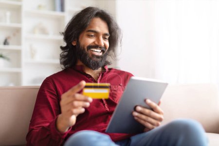 Smiling Indian man holding a credit card and a digital tablet, likely shopping online from home, copy space