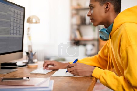 Téléchargez les photos : Adolescent gars en sweat à capuche jaune se concentrant sur le travail à son bureau d'ordinateur, prenant des notes avec stylo bleu dans un espace d'étude lumineux et organisé - en image libre de droit