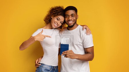 Téléchargez les photos : Couple afro-américain souriant tenant des passeports et pointant du doigt, suggérant des plans de voyage sur un fond jaune - en image libre de droit