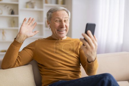 Happy senior male using a smartphone to video chat, enthusiastically waving at the screen with a joyous expression