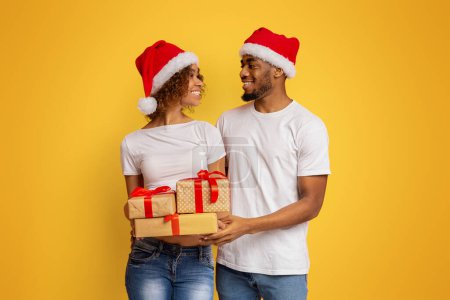Photo for Happy couple ready for Christmas holidays. African-american man and woman standing with lots of presents, smiling to each other, orange background - Royalty Free Image