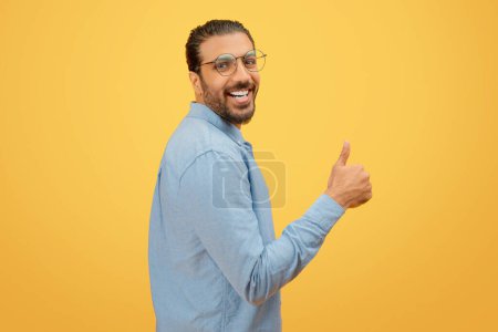 Positive indian man with a beard and glasses showing thumbs up against a yellow background