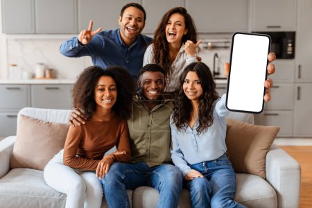 Foto de Grupo de amigos diversos posando en un sofá con uno sosteniendo un teléfono inteligente con una pantalla en blanco, sonriendo a la cámara - Imagen libre de derechos