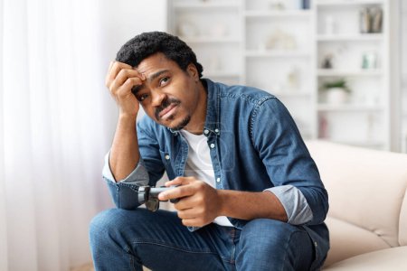 Téléchargez les photos : Un jeune homme noir assis sur le canapé, l'air inquiet tout en tenant un joystick, jouer à des jeux vidéo à la maison dans une pièce bien éclairée et toucher la tête - en image libre de droit