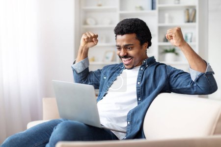 Téléchargez les photos : Un homme noir enchanté célébrant le succès avec un ordinateur portable à la maison, assis sur un canapé lève joyeusement les poings dans un geste de victoire tout en regardant l'écran d'ordinateur - en image libre de droit