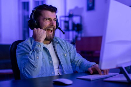 Gamer bearded guy in a denim shirt talks into his headsets microphone while engaged in a game, celebrating win