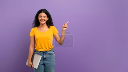Foto de Mujer sonriente en la parte superior amarilla apuntando hacia arriba y sosteniendo una tableta digital, sobre un fondo púrpura - Imagen libre de derechos