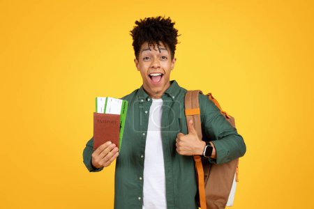 Enthusiastischer junger schwarzer Reisender hält einen Pass und zeigt den Daumen nach oben, symbolisiert die Zustimmung zu Reiseabenteuern auf gelbem Hintergrund