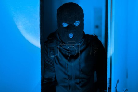 Photo for A shadowy man masked burglar stands in a doorframe, bathed in a surreal blue light, robing apartment - Royalty Free Image