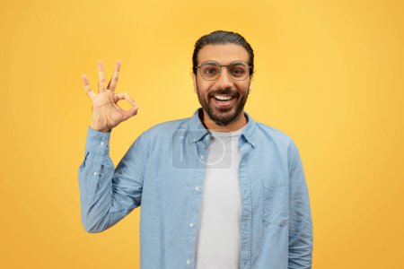 Sonriente hombre oriental barbudo en gafas dando el signo OK con la mano sobre un fondo amarillo