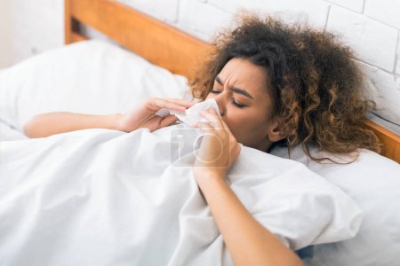 Foto de Mujer joven enferma sonándose la nariz, teniendo gripe o resfriado, acostada en la cama - Imagen libre de derechos