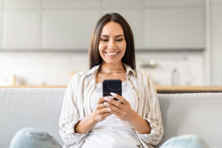 Téléchargez les photos : Une femme confiante interagit avec son smartphone, rayonnant de positivité à partir d'un cadre confortable salon - en image libre de droit