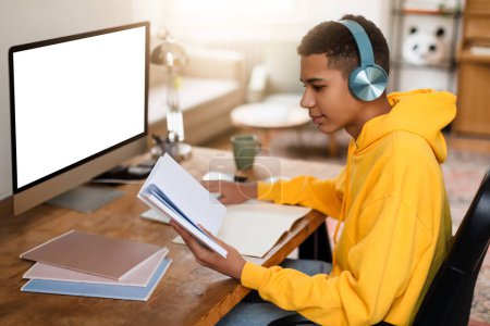 Foto de Un joven enfocado en una sudadera con capucha amarilla estudia desde un cuaderno con una computadora y auriculares en su escritorio - Imagen libre de derechos