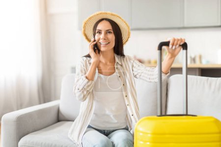 Photo for Joyous lady traveler on phone, with a suitcase and passport, planning her trip while sitting on a sofa, calling taxi - Royalty Free Image