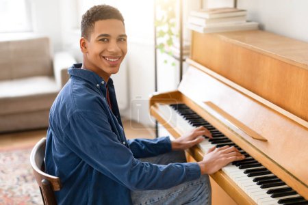 Photo for Happy black teenager guy wearing blue shirt sitting at piano, smiling as he looking and smiling at camera while playing, filled with joy in cozy home setting - Royalty Free Image