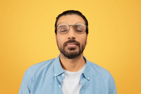 Ruhiger und entspannter indischer Mann mit Bart, der die Augen schließt, Brille auf gelbem Hintergrund