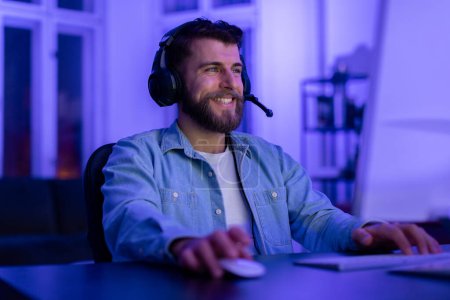 Photo for Cheerful young bearded man wearing headset smiles while engaged in an online game late at night at home - Royalty Free Image