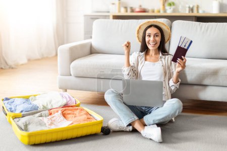 Happy woman traveler with laptop showcases passport and boarding pass, ready for a journey, sitting on the floor