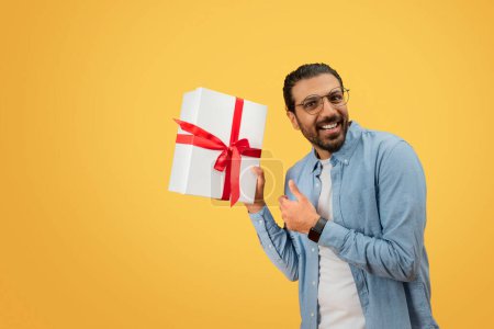 An elated indian man in a casual denim shirt presents a white gift box with a red ribbon, set against a yellow background