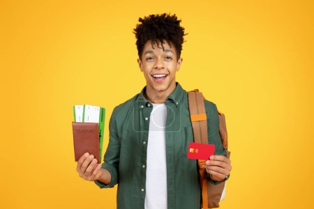 A self-assured young african american guy wearing a backpack holds a passport and credit card, representing readiness for travel on yellow