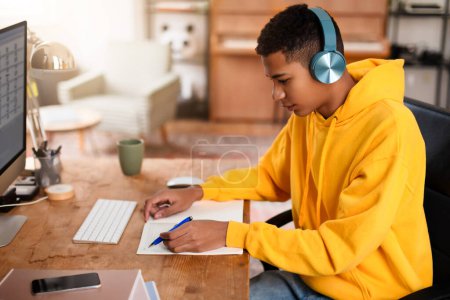 Foto de Una persona se ve escribiendo en un cuaderno en su escritorio, con una computadora y auriculares cerca - Imagen libre de derechos