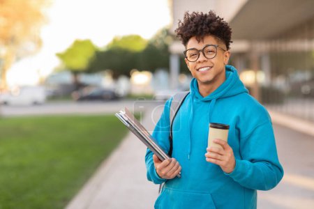 Ein fröhlicher brasilianischer Student mit Brille hält eine Tasse Kaffee und Ordner in der Hand und posiert draußen in der Stadt