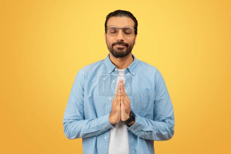 Photo for Adult indian guy in a blue shirt stands with hands in a prayer pose and eyes closed against a yellow background - Royalty Free Image