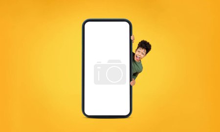 Lächelnder junger afrikanisch-amerikanischer Mann, der sich hinter einem riesigen Smartphone mit leerem Bildschirm auf gelbem Hintergrund versteckt